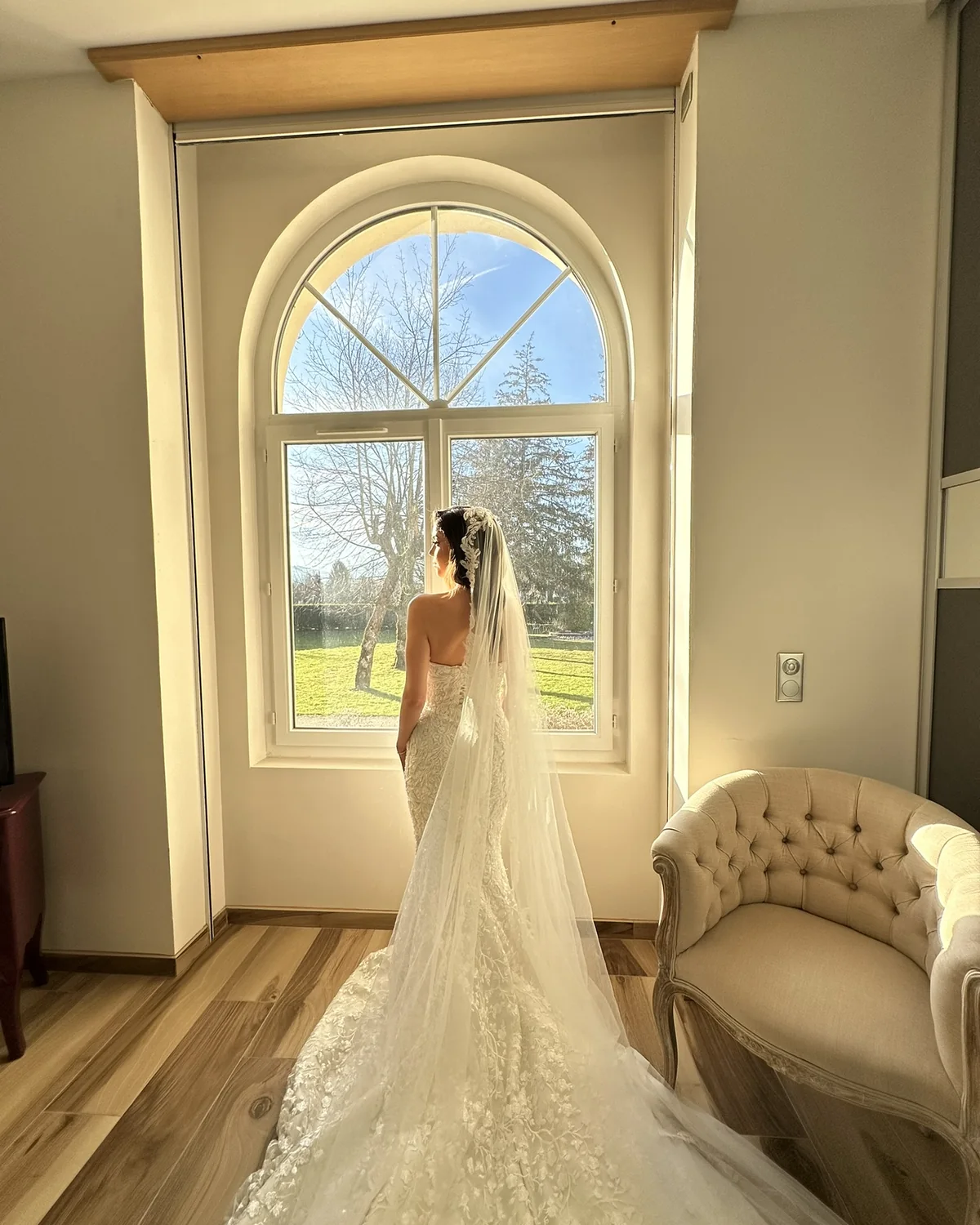 Une mariee de dos, robe a traine et long voile, devant une fenetre cintree