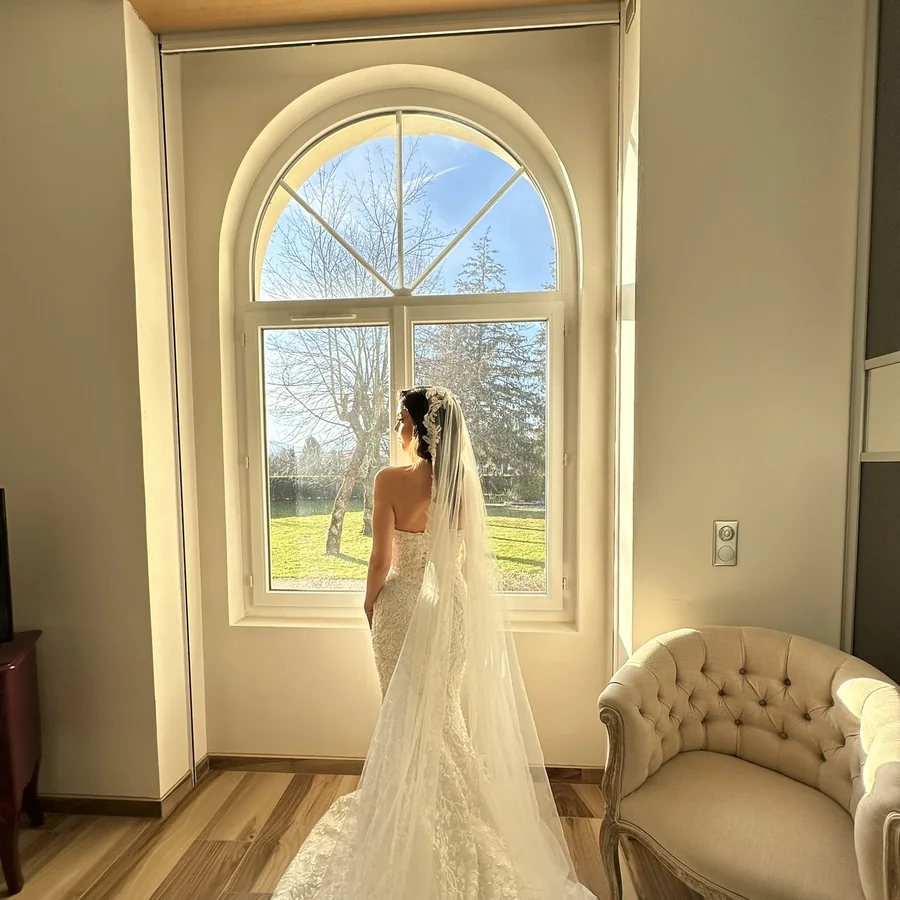 Une mariee de dos, en robe et voile, devant une fenetre cintree